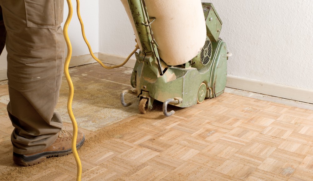 Expert Wood Floor Sanding in Hounslow—technician using a green drum sander to refinish an oak parquet floor, exposing clean timber beside worn surface.