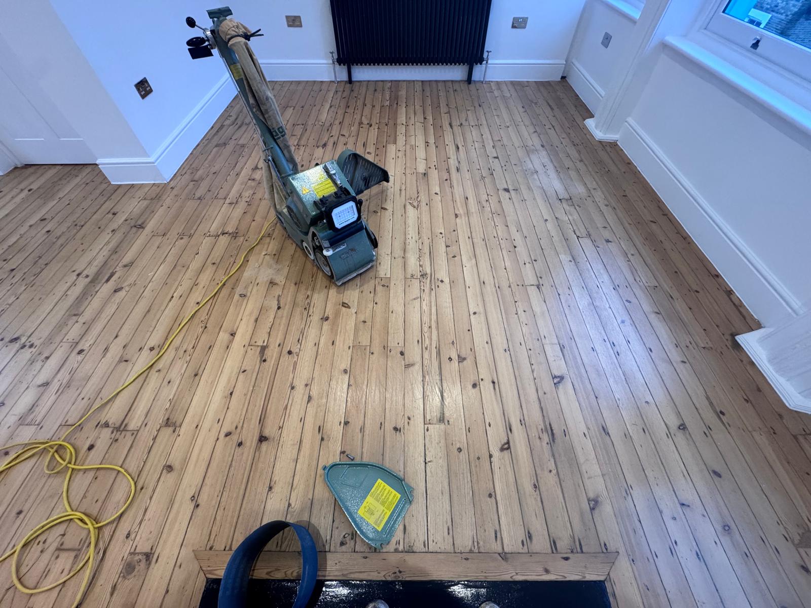 Wood Floor Sanding in Hounslow on narrow pine softwood floorboards in a living room, with a professional belt sander preparing the timber for a fresh finish.